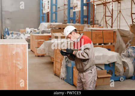 Der Lagerhalter, markiert die Ankunft auf dem Tablett. Mann, der an einem Gabelstapler durch ein Lager in einer Fabrik. Treiber in Uniform und Schutzhelm. Das Konzept der Logistik und Lagerung Stockfoto