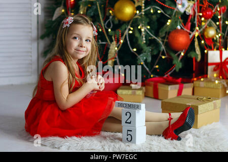 Llittle Mädchen im roten Kleid sitzt auf dem Teppich. Würfel mit Inschrift am 25. Dezember. Weihnachtsbaum mit Ornamenten im Hintergrund. Happy childh Stockfoto