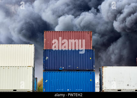 Liefer- Containern bei der Lagerung mit Federn des schwarzen toxischen Rauch gestapelt aus einem Feuer hinter. Stockfoto