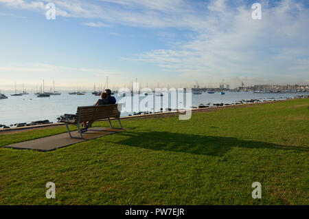 Die Silhouette eines Paar auf einer Bank sitzen durch das Wasser die Sonne genießen und Vista von Wasser und Boote Stockfoto