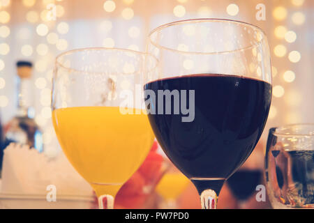 Nahaufnahme der Gläser mit Rotwein und Orangensaft auf einem Tisch im Restaurant auf die Girlanden Vorhang Hintergrund. Gefüllte Gläser in einem Restaurant serviert. Stockfoto