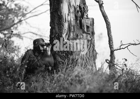 Versteckte unbekannte Re-Enactor als Deutsche Wehrmacht Infanterie Soldat im Zweiten Weltkrieg Soldat sitzt im Hinterhalt in Wald und Suchen an der alten Arm gekleidet Stockfoto