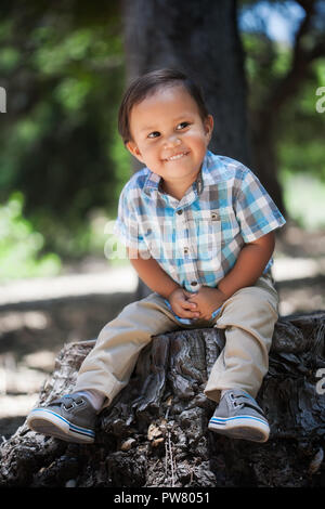 Latino Jungen biss sich auf die Lippe mit einem netten Lächeln, sitzen auf einem Baumstumpf in der freien Natur seine Hände greifen mit Plaid Shirt Stockfoto