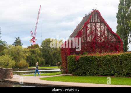 Herbst Blick auf die junge Frau zu Fuß & mit Telefon von See & lych Gate - wunderschöne, friedliche, kommunale Rowntree Memorial Park, York, England, UK. Stockfoto