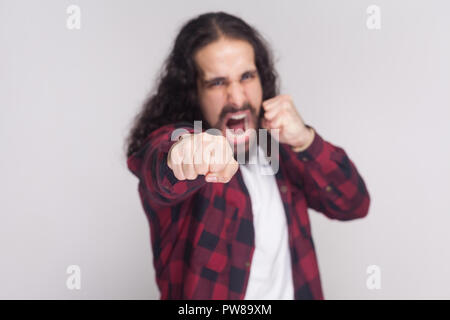 Wütend schreienden Mann mit Bart und den schwarzen langen Locken in der beiläufigen kariert rot Shirt stehend, auf Kamera und Angriff mit Boxing Fäuste. Ich Stockfoto