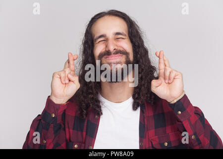 Portrait von hoffnungsvollen stattlicher Mann mit Bart und schwarzen langen Locken im lässigen Stil, kariert rot Shirt ständigen geschlossenen Augen mit gekreuzten Fingern. Stockfoto