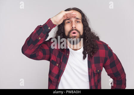 Portrait von überrascht Mann stattlicher Mann mit Bart und schwarzen langen Locken in der beiläufigen kariert rot Shirt stehen und weit schauen mit entsetzten Gesicht. Ich Stockfoto