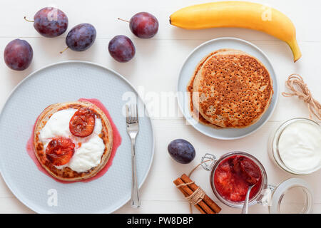 Weiße Holztisch mit closeup Bild von Dessert Untertasse mit Gipfeli, Pfannkuchen, saure Sahne und frischen Pflaumen. Das Glas mit gekochten Pflaumen, frisch Pflaumen Stockfoto