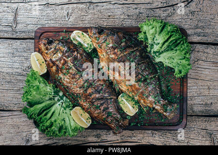 Ein köstlich Gebratener Karpfen auf einem Grill, auf einem Holzbrett vorgestellt, und entlang der Blätter von grüner Salat und Stücke von Zitrone. Stockfoto