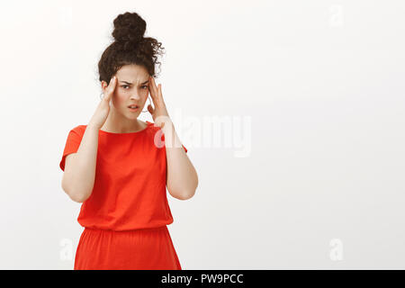 Portrait von unfocused unruhigen kaukasische Mädchen im roten Kleid,, Hände auf Tempel und runzelte die Stirn von Unmut, leiden unter Kopfschmerzen oder Migräne, abbrechen Pläne von schmerzliche Gefühl Stockfoto