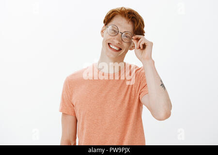 Freut mich. Smart attraktive rothaarige Menschen mit Sommersprossen und Tätowierungen, berühren Rim von Brillen, Neigekopf, lächelnd mit freundlich und froh, Ausdruck, neue Leute treffen in der Universität Stockfoto
