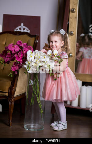 Blue-eyed niedliche Mädchen in ein rosa Kleid Holding eine Vase mit Orchideen und lächelnd Stockfoto