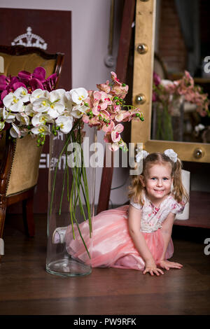 Blue-eyed niedliche Mädchen in ein rosa Kleid sitzt in der Nähe von eine Vase mit Orchideen und lächelnd Stockfoto