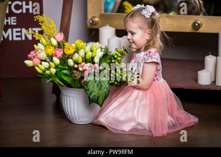 Blue-eyed süßen Mädchen in ein rosa Kleid sitzt in der Nähe von eine Vase mit Tulpen, Mimose, Beeren und Grüns und lächelnd Stockfoto