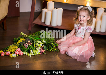 Blue-eyed niedliche Mädchen in ein rosa Kleid sitzt auf dem Boden neben einem Blumenstrauß aus Tulpen, Mimose, Beeren und Grün Stockfoto