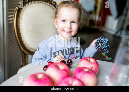 Blue-eyed niedliche Mädchen an einem Tisch sitzen mit Äpfeln, Kirschen, Trauben und lächelnd Stockfoto