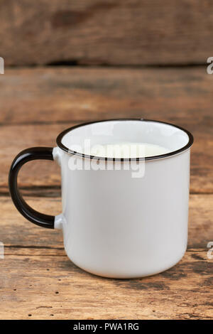 Emaille Tasse Milch auf Holz- Hintergrund. Frische Milch in eine weiße Tasse Emaille auf rustikalen hölzernen Brettern, vertikale Bild. Stockfoto