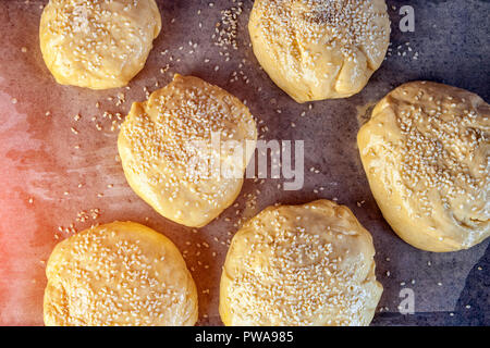 Eine Nahaufnahme der Prozess des Kochens Brötchen Mit Sesam oder Pfannkuchen auf einen hölzernen Tisch. Flach: Hamburger Brötchen auf Pergament Papier bereit zum Kochen Stockfoto