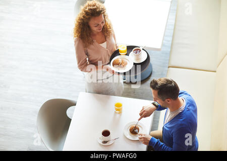 Junge Kellnerin holding Fach mit frischen Pfannkuchen, Tasse Kaffee und ein Glas Orangensaft im Stehen durch die Tabelle, in der Geschäftsmann sein Frühstück essen Stockfoto