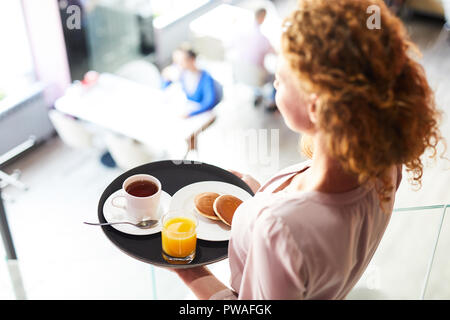 Junge Frau, die Pfannkuchen, Kaffee und Saft auf Fach während der Bewegung zu einem der Tische im Cafe Stockfoto