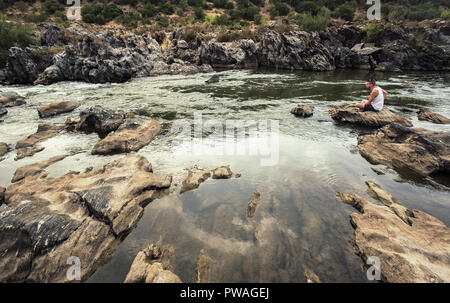 Junger Mann in kurzen weißen Hemd sitzt nachdenklich auf den Felsen des Guadiana Stockfoto