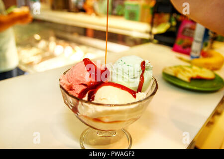 Weibliche Verkäufer gießt Soße auf Eis. Frau schwer machen Eis Kupfer Schale mit Obst und Süßigkeiten schmilzt in ein hohes Becherglas. Stockfoto