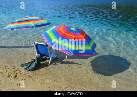 Sommer Zubehör auf einem Strand, zwei bunte Sonnenschirme, blau Liege, schnorcheln Maske und Flossen im klaren, seichten Wasser des Ionischen Meeres Stockfoto