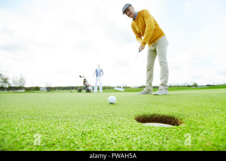 Reifer Mann in casualwear stehen auf der grünen Wiese und gehen Golf Ball in das Loch zu schlagen Stockfoto