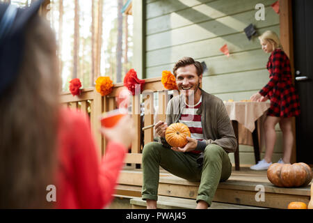 Junger Mann mit Kürbis sitzen auf der Terrasse und im Gespräch mit kleinen Tochter auf dem Hintergrund seiner Frau mit Tisch für das Mittagessen Stockfoto