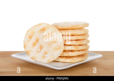 Stapel der Japanische Reiskräcker auf einem kleinen weißen Quadrat Porzellanteller auf Holz Tisch isoliert hinter sich. Reis Cracker sind in der Regel niedrig in Zucker und Fett. Stockfoto