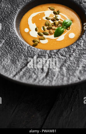 Nahaufnahme von Kürbis und Karotten Suppe mit Kürbiskernen, Sahne und Basilikum in Schwarz moderne Platte auf dunklen Holztisch mit kopieren. Ansicht von oben. Stockfoto