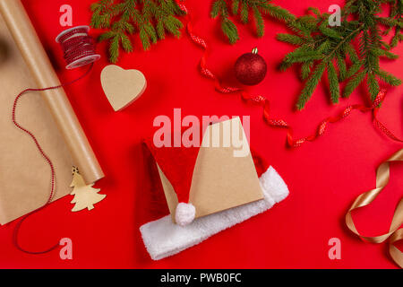 Vorbereitung für Weihnachten: Geschenkpapier, Dekorationen und Gegenwart Geschenkkarton mit Santa's Hut auf rotem Hintergrund Stockfoto