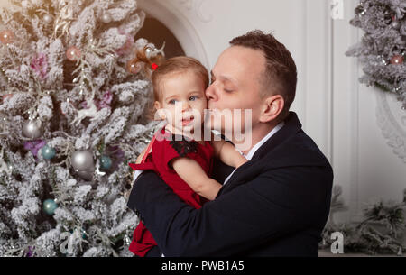 Frohe Weihnachten und guten Rutsch ins neue Jahr. Vati in einen Anzug küssen, seine Tochter in einem eleganten roten Kleid auf dem Hintergrund der Weihnachtsbaum gekleidet Stockfoto