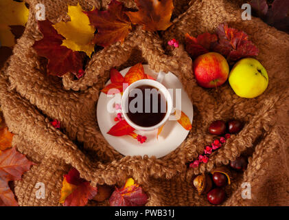 Weiße Tasse mit heißem Kaffee, Dampf, Wollstoff umgibt eine Untertasse, bunten Ahornblätter auf einem Tuch, zwei Äpfeln und Kastanien, ein Herbst, Stockfoto