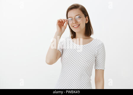 Charmante kaukasische Mädchen in trendigen t-shirt, das Berühren der Rand der Gläser und breit grinsend, im Gespräch mit Kollegen und diskutieren zukünftige Ereignis, lässig stehend über grauer Hintergrund, Kühlen Stockfoto