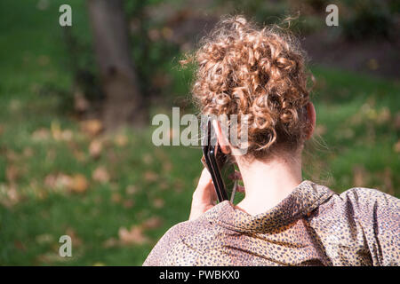 Eine rothaarige Frau, die allein auf einer Parkbank sitzt und einen Anruf auf ihrem Handy in London macht Stockfoto