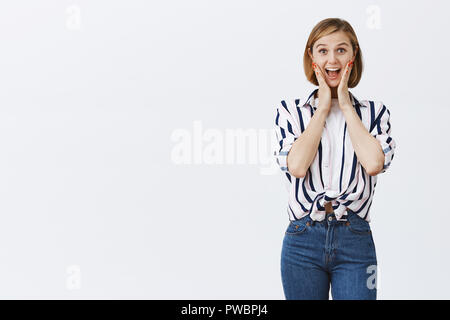 Frau Screams von Unterhaltung und Freude, die erstaunt. Portrait von attraktiven aktiv und glückliche junge weibliche Mitarbeiter in stilvollen gestreifte Bluse, t-shirt, holding Palmen auf Wangen, lächelnd und Schreien Stockfoto