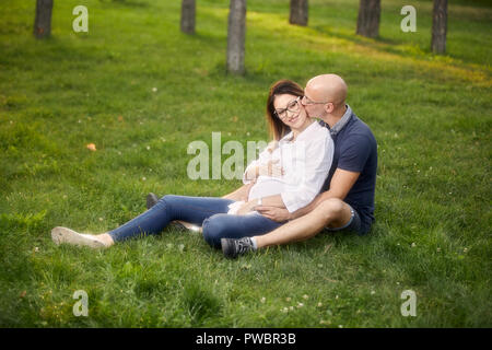 Junges Paar sitzt im Gras Feld in Park. lächelt glücklich, Mann küssen schwangere Frau. Stockfoto