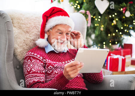 Ein Porträt der ältere Mann mit Santa Hut auf dem Sessel zu Hause an Weihnachten sitzen, mit Tablette. Stockfoto