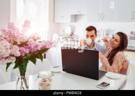Glückliches junges Paar mit Laptop beim Frühstück in der modernen Küche. Junger Mann und Frau trinken Kaffee und Lächeln. Fokus auf Laptop Stockfoto