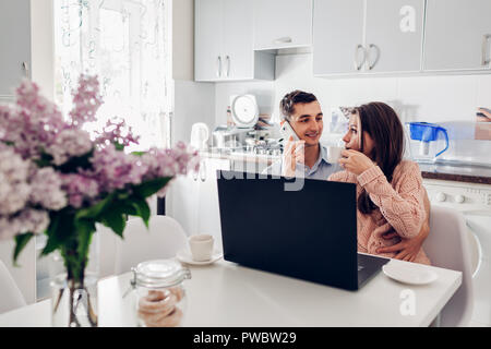 Glückliches junges Paar mit Laptop beim Frühstück in der modernen Küche. Junger Mann am Telefon sprechen, während Frau trinkt Kaffee Stockfoto