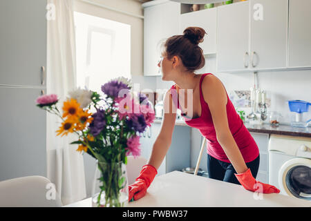 Junge Frau in Handschuhe Reinigung der Oberfläche mit Schwamm in der modernen Küche mit Blumen geschmückt. Stockfoto