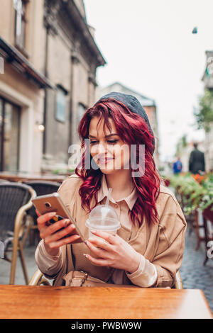 Schöne junge Frau mit Kaffee im Café im Freien, während Sie ihr Smartphone. Portrait von stilvollen Mädchen soziales Netzwerk prüfen Stockfoto