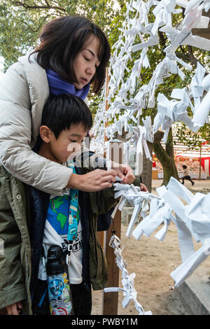 Japanische neues Jahr, shogatsu. Junge Frau, Kind, Junge, 5-6 Jahre, schlechtes Vermögen Papierstreifen zu binden, Omikuji zu Rahmen an der Nishinomiya Schrein. Stockfoto