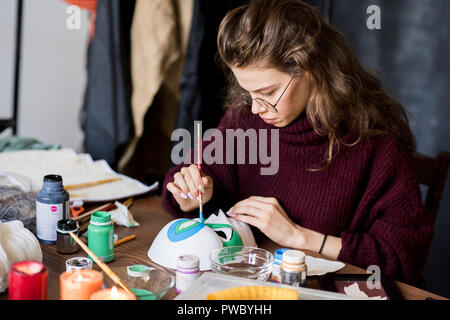 Ernsthafte qualifizierte attraktive junge Frau in Gläsern am Tisch sitzen und die Fokussierung auf die Arbeit beim Malen Maske mit Pinsel in der eigenen Art Studio Stockfoto