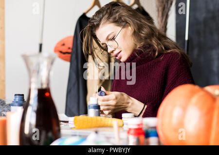 Positive attraktive junge Frau in Gläsern am Tisch sitzen und lesen Label auf Paint jar während der Vorbereitung für das Mischen von Farben in art studio Stockfoto