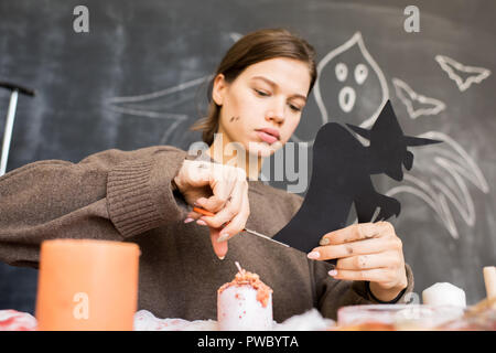Ernsthafte beschäftigt Mädchen in Pullover sitzen am Tisch gegen Tafel mit Ghost und schneiden Hexe aus schwarzem Papier während der Arbeit an Halloween Deko Stockfoto