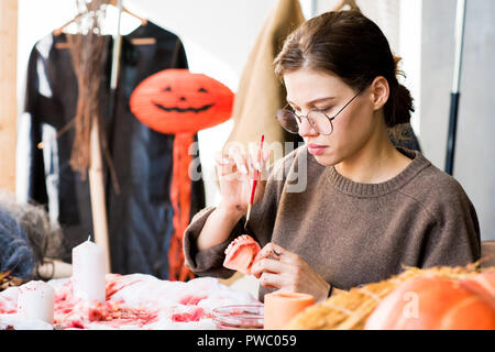 Ernsthafte schöne junge Dame in Gläsern am Tisch voll von Halloween und Malerei Spielzeug Kiefer sitzend mit Farbe von Blut Farbe beim Arbeiten auf costu Stockfoto