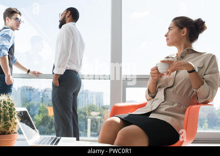 Schweren Träumen junge geschäftsfrau mit Haar bun Sitzen im Stuhl im Büro und trinkt Kaffee und multi-ethnischen Unternehmer, die miteinander sprechen, Stockfoto