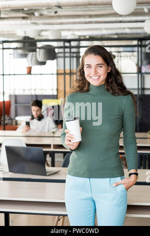 Glücklich, aufgeregt, junge Frau mit dem lockigen Haar stand im Büro mit modernem Design und Trinken takeout Kaffee, während ein Lächeln auf die Kamera Stockfoto